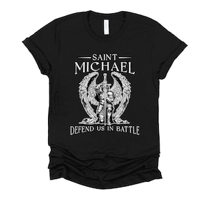 May include: Black t-shirt featuring a detailed graphic of Saint Michael, a winged figure holding a sword. The text above reads "SAINT MICHAEL," and below, "DEFEND US IN BATTLE." The design is in white, contrasting against the black fabric.