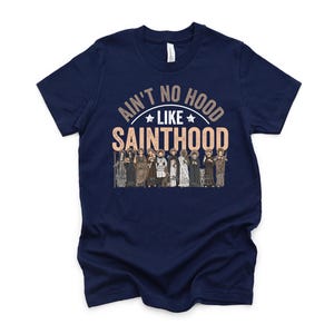 Pu&ograve; includere: T-shirt blu navy con la scritta "Ain't No Hood Like Sainthood" in un design circolare. Sotto il testo c'&egrave; una grafica di diverse figure in abiti religiosi. La maglietta ha un girocollo.