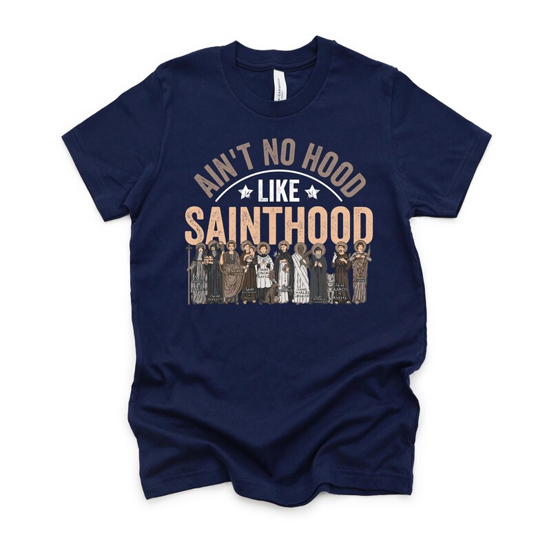 Pu&ograve; includere: T-shirt blu navy con la scritta "Ain't No Hood Like Sainthood" in un design circolare. Sotto il testo c'&egrave; una grafica di diverse figure in abiti religiosi. La maglietta ha un girocollo.
