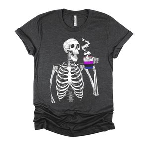 May include: A dark heather grey t-shirt with a skeleton holding a coffee mug with the bisexual pride flag colors. The skeleton is smiling and has a steam coming from the mug.