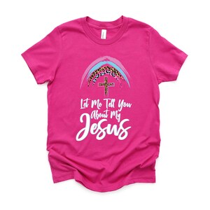May include: Pink t-shirt with a rainbow and cross graphic. The rainbow is made of blue, pink, and white stripes with a leopard print design. The text on the shirt reads "Let Me Tell You About My Jesus".
