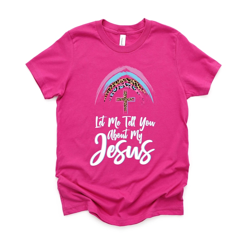May include: Pink t-shirt with a rainbow and cross graphic. The rainbow is made of blue, pink, and white stripes with a leopard print design. The text on the shirt reads "Let Me Tell You About My Jesus".