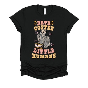 May include: Black t-shirt with a graphic design featuring a skeleton holding a coffee cup. The text reads "DATA COFFEE AND LITTLE HUMANS" in a retro font with pink and yellow accents. The design includes stars and a coffee cup.