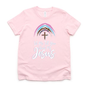 May include: Pink t-shirt with a rainbow design featuring a cross and leopard print. The text "Let Me Tell You About My Jesus" is printed on the shirt.