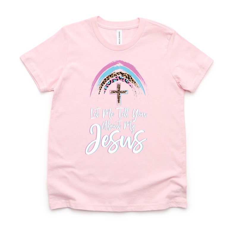 May include: Pink t-shirt with a rainbow design featuring a cross and leopard print. The text "Let Me Tell You About My Jesus" is printed on the shirt.