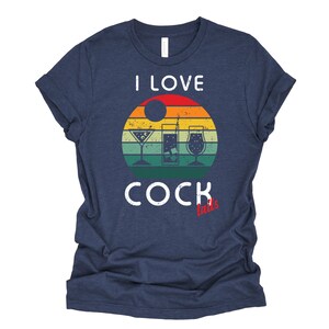Op de afbeelding: Een marineblauw t-shirt met een vintage-stijl grafische afbeelding met de tekst "Ik hou van cocktails" met een zonsondergang achtergrond en drie cocktailglazen.