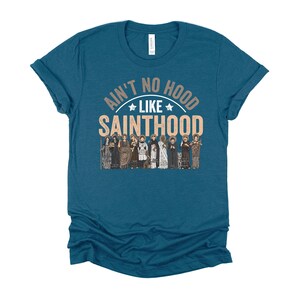 Pu&ograve; includere: T-shirt color teal con la scritta "Ain't No Hood Like Sainthood" in un design circolare, con un gruppo di figure sottostante. La maglietta &egrave; a tinta unita e ha maniche corte.