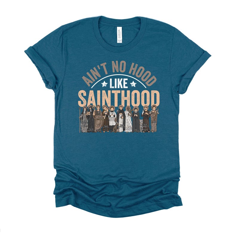 Pu&ograve; includere: T-shirt color teal con la scritta "Ain't No Hood Like Sainthood" in un design circolare, con un gruppo di figure sottostante. La maglietta &egrave; a tinta unita e ha maniche corte.