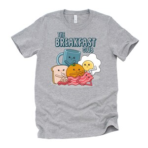 May include: A heather grey t-shirt featuring a cartoon illustration of breakfast foods. The text "The Breakfast Club" is printed on the shirt.