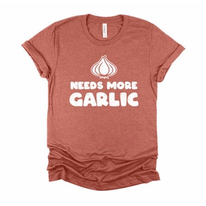 May include: A rust-colored t-shirt with a white graphic of a garlic clove and the text "Needs More Garlic".