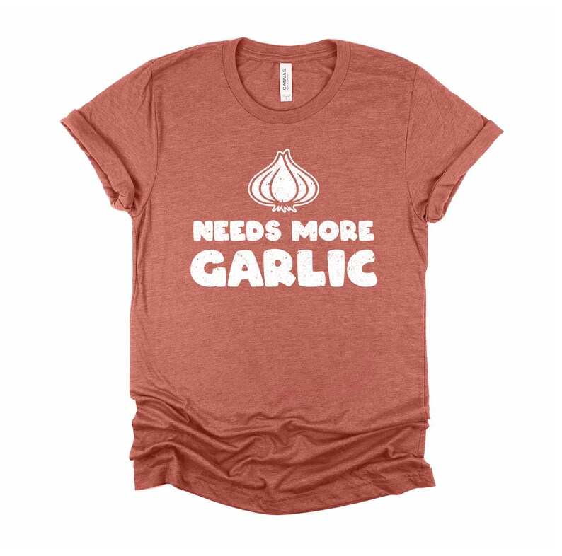 May include: A rust-colored t-shirt with a white graphic of a garlic clove and the text "Needs More Garlic".