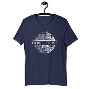 May include: Navy blue t-shirt with a white graphic of a clock face made of gears and the word "HOROLOGIST" printed in white.