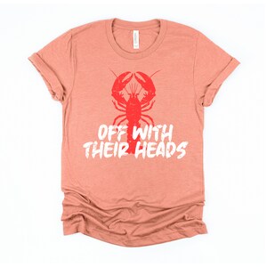 K&ouml;nnte beinhalten: Ein korallenfarbenes T-Shirt mit einem roten Hummer-Motiv und dem Text "Off with their heads" in Wei&szlig;.