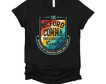 May include: Black t-shirt with a colourful graphic. The design features the text "The Oxford Comma Preservation Society" in a retro style. The graphic includes a shield shape and the words "Defenders of Tradition, Form, and Clarity."