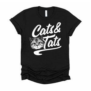 May include: Black t-shirt with a white graphic of a cat's face and the text "Cats & Tats" in a retro font.