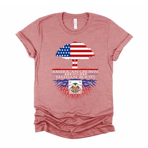 May include: A heather red t-shirt with a graphic of a tree with the American flag as the leaves and the Haitian flag as the roots. The text "AMERICAN GROWN WITH HAITIAN ROOTS" is printed on the shirt.