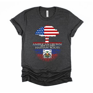 May include: A dark heather grey t-shirt with a tree design. The tree's trunk is formed by the Haitian flag, and the tree's crown is formed by the American flag. The text "AMERICAN GROWN WITH HAITIAN ROOTS" is printed on the shirt.
