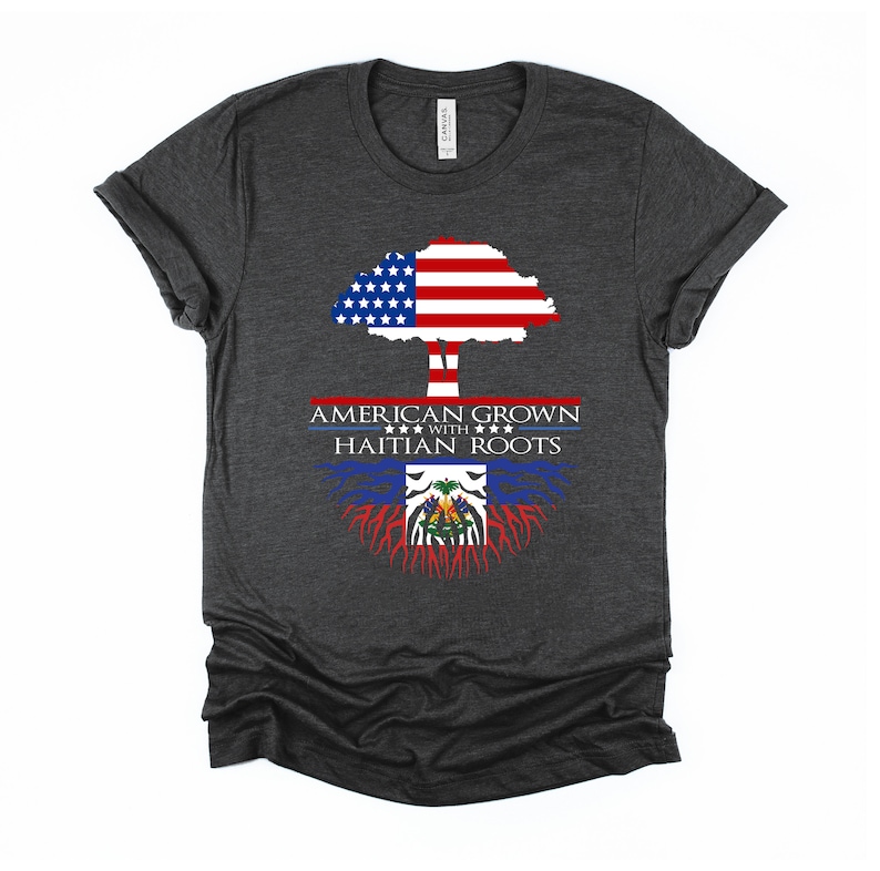 May include: A dark heather grey t-shirt with a tree design. The tree's trunk is formed by the Haitian flag, and the tree's crown is formed by the American flag. The text "AMERICAN GROWN WITH HAITIAN ROOTS" is printed on the shirt.