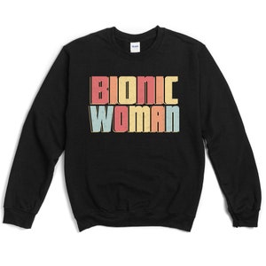 May include: Black sweatshirt with the text "BIONIC WOMAN" in a retro font. The text is in a layered design with red, yellow, and blue colors. The sweatshirt is a crew neck style.