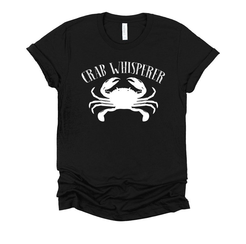 May include: Black t-shirt with white text that reads "Crab Whisperer" and a white silhouette of a crab.