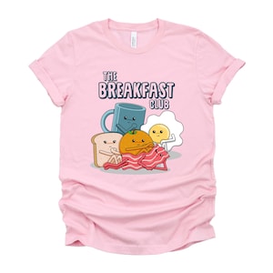 May include: Pink t-shirt featuring a graphic of breakfast foods, including a mug, orange, toast, bacon, and egg. The text "The Breakfast Club" is printed on the shirt.