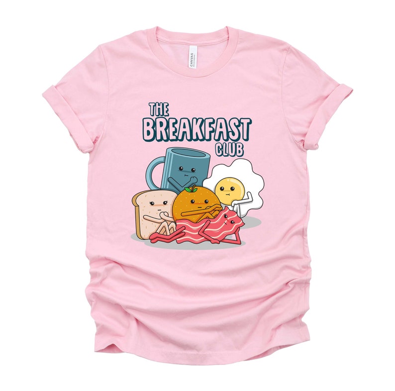 May include: Pink t-shirt featuring a graphic of breakfast foods, including a mug, orange, toast, bacon, and egg. The text "The Breakfast Club" is printed on the shirt.