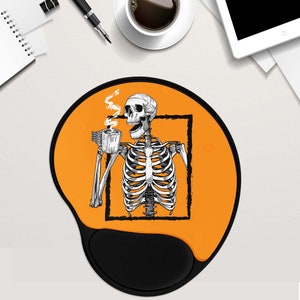 May include: Orange and black mouse mat with a skeleton holding a cup of coffee. The skeleton is inside a black square frame. The mouse mat has a black, ergonomic wrist rest.