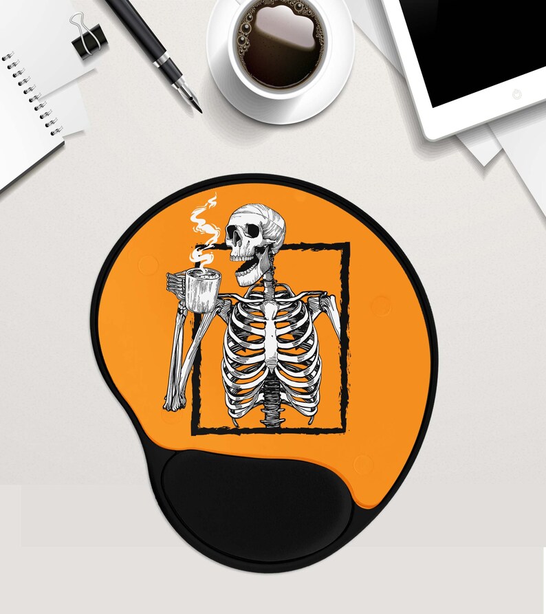 May include: Orange and black mouse mat with a skeleton holding a cup of coffee. The skeleton is inside a black square frame. The mouse mat has a black, ergonomic wrist rest.