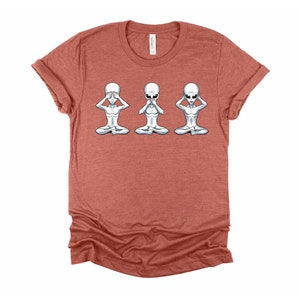 May include: A heather red t-shirt featuring a graphic of three grey aliens in a row. The aliens are in a yoga pose, with one covering their eyes, one covering their mouth, and one covering their ears.