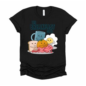 May include: Black t-shirt featuring a graphic of breakfast foods, including a cup of coffee, an orange, a slice of toast, bacon, and an egg. The text "The Breakfast Club" is printed above the graphic.