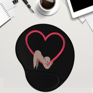 May include: Black mouse pad with a pink heart and a brown sloth hanging from it. The sloth is holding onto the heart with its paws.