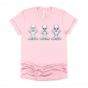 May include: Pink t-shirt featuring a graphic of three grey aliens in a row, each with one hand covering their eyes, one hand covering their ears, and one hand covering their mouth. The aliens are sitting in a yoga pose.