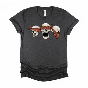 May include: A dark heather grey t-shirt with a graphic of three skulls wearing red bandanas. The skulls are blindfolded with the bandanas.
