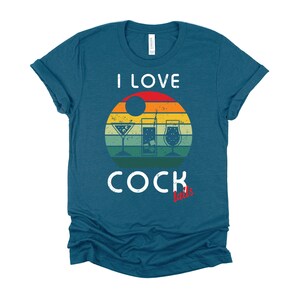 Op de afbeelding: Een tealblauw t-shirt met een vintage-stijl grafische afbeelding van een zonsondergang met drie cocktailglazen. De tekst "Ik hou van cocktails" is op het shirt gedrukt.