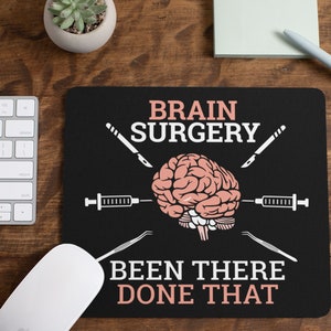 May include: Black mouse pad with white text that reads "Brain Surgery Been There Done That". The text is surrounded by a brain graphic, a scalpel, and a syringe.