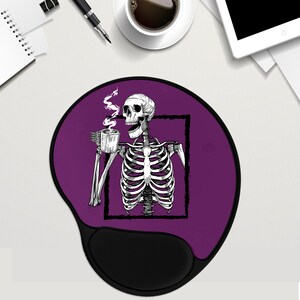 May include: Purple mouse mat with a black skeleton holding a cup of coffee. The skeleton is framed in a black square. The mouse mat has a black, ergonomic wrist rest.