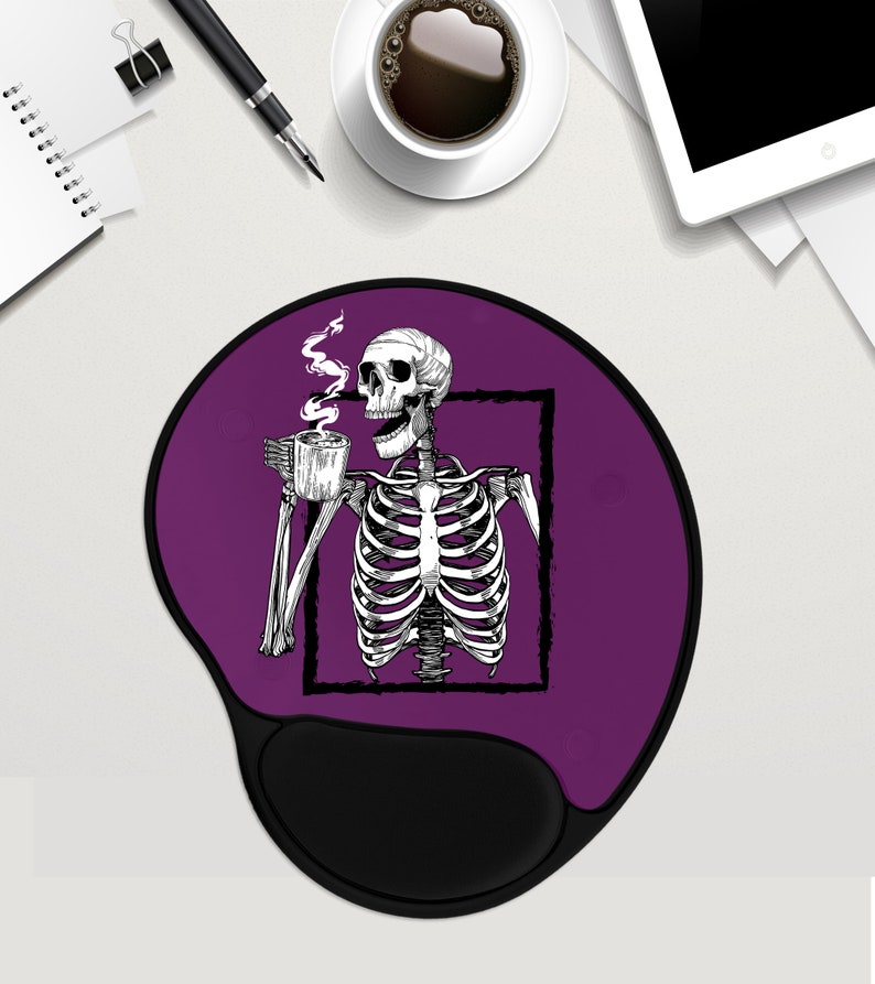 May include: Purple mouse mat with a black skeleton holding a cup of coffee. The skeleton is framed in a black square. The mouse mat has a black, ergonomic wrist rest.