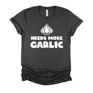 May include: A dark heather grey t-shirt with a white graphic of a garlic bulb and the text "Needs More Garlic".