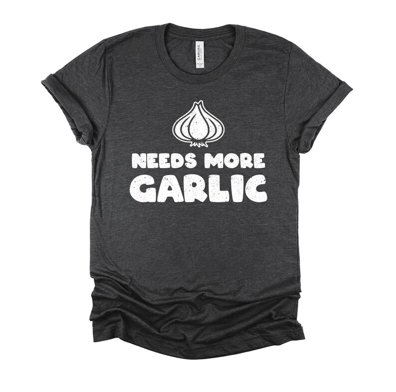 May include: A dark heather grey t-shirt with a white graphic of a garlic bulb and the text "Needs More Garlic".