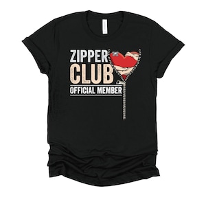 May include: Black t-shirt with the text "ZIPPER CLUB OFFICIAL MEMBER" in white and tan. A red heart with bandages is featured, with a zipper design. The shirt is made of a soft material.