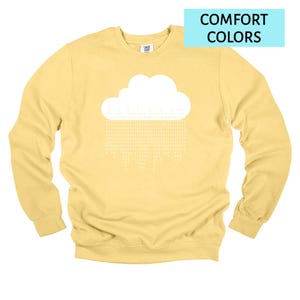Op de afbeelding: Een gele sweatshirt met een witte wolkengrafiek en de tekst "COMFORT COLORS" in een blauwe rechthoek. Het wolkenontwerp heeft een regenachtig patroon van witte tekst. De sweatshirt is een crew neck stijl.