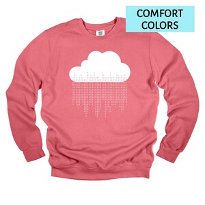 Op de afbeelding: Een koraalkleurige sweatshirt met een witte wolkengrafiek en binaire code regen. De tekst "COMFORT COLORS" staat in een lichtblauwe rechthoek. De sweatshirt heeft lange mouwen en een ronde hals.