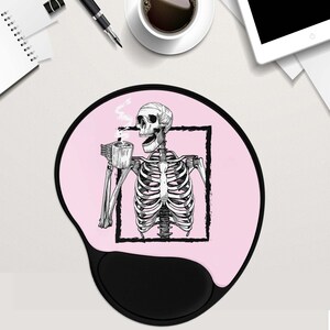 May include: Pink mouse mat with a black skeleton holding a cup of coffee. The skeleton is framed in a black square. The mouse mat has a black, ergonomic wrist rest.