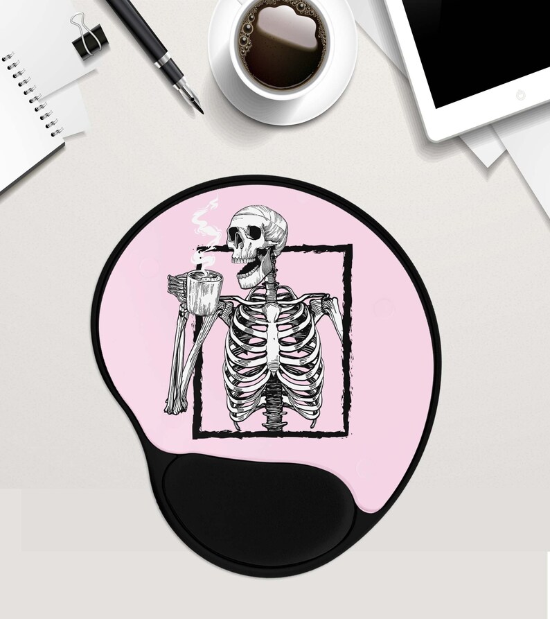 May include: Pink mouse mat with a black skeleton holding a cup of coffee. The skeleton is framed in a black square. The mouse mat has a black, ergonomic wrist rest.