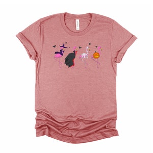 May include: A pink heather t-shirt with a graphic of four pink flamingos dressed in Halloween costumes. The flamingos are dressed as a witch, a vampire, a mummy, and a pumpkin.