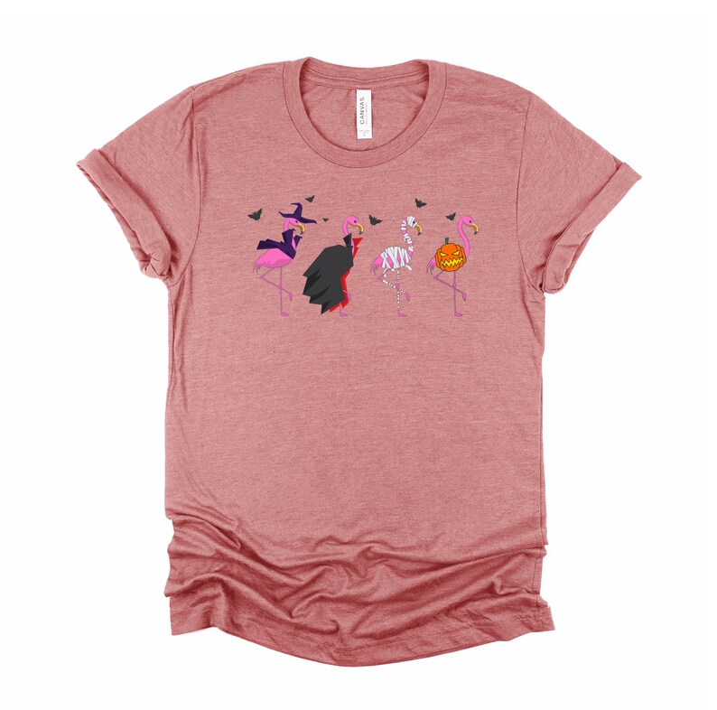 May include: A pink heather t-shirt with a graphic of four pink flamingos dressed in Halloween costumes. The flamingos are dressed as a witch, a vampire, a mummy, and a pumpkin.