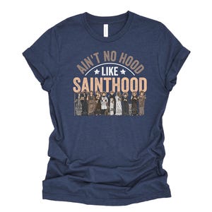 Pu&ograve; includere: T-shirt blu navy con la scritta "Ain't No Hood Like Sainthood" in marrone e bianco. La grafica include illustrazioni di varie figure. La maglietta &egrave; realizzata in un materiale morbido.