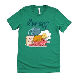 May include: A green t-shirt featuring a cartoon illustration of breakfast foods. The text "The Breakfast Club" is printed on the shirt.