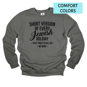 May include: A dark gray sweatshirt with long sleeves. The front of the sweatshirt features the text "SHORT VERSION OF EVERY Jewish HOLIDAY • THEY TRIED TO KILL US • WE WON." A light blue rectangle in the upper right corner reads "COMFORT COLORS."