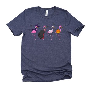 May include: A navy blue t-shirt with a graphic of five pink flamingos dressed in Halloween costumes. The flamingos are wearing a witch hat, a vampire cape, a mummy costume, a skeleton costume, and a pumpkin costume.